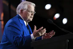 A rotund grey-haired man in a blue suit