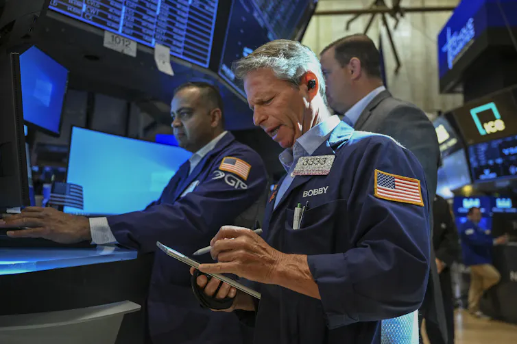 People in ties and stock-trader jackets stand near multiple electronic monitors.