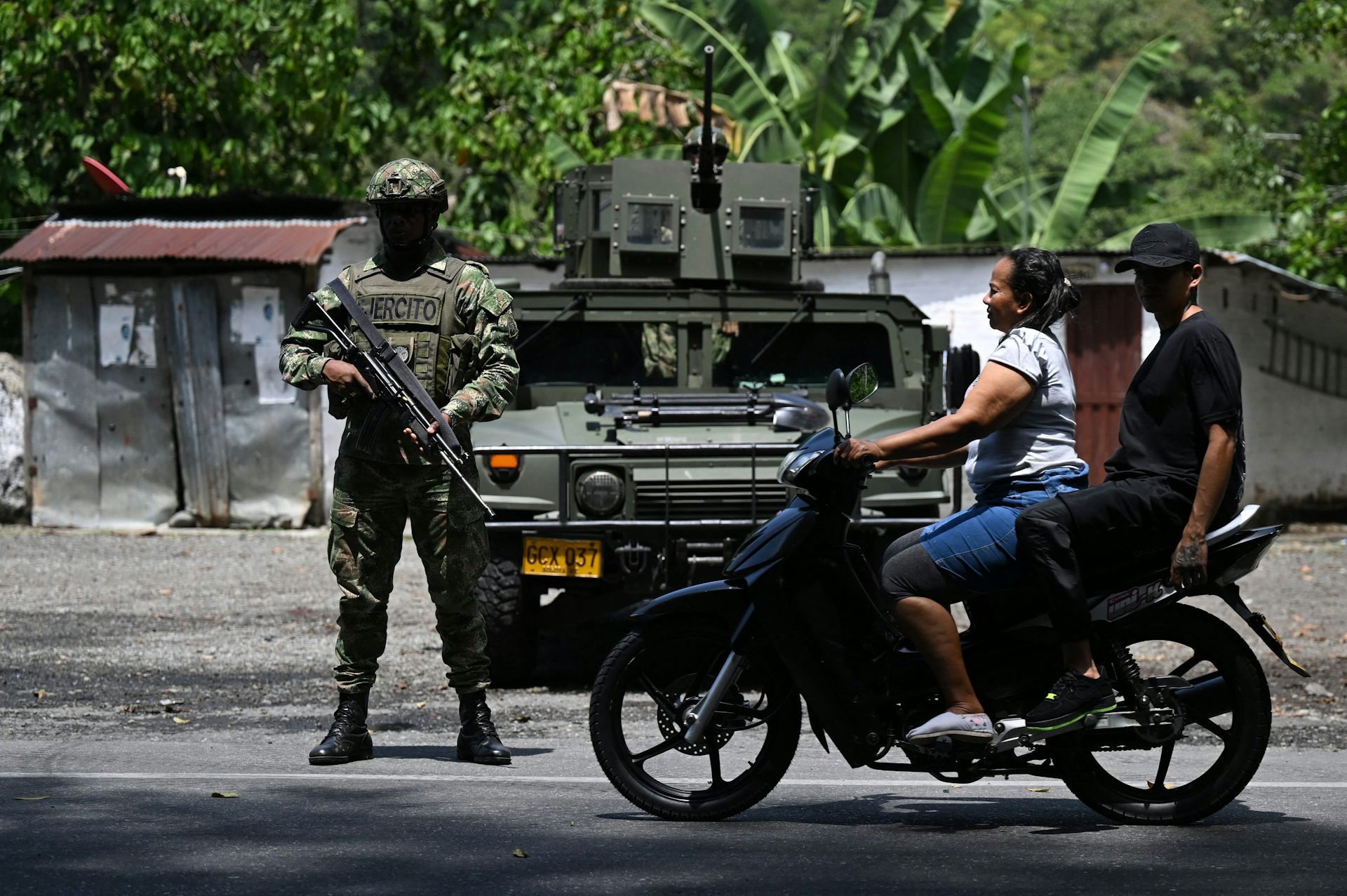 Duas pessoas em uma motocicleta passam por um homem em uniforme militar que carrega uma arma grande.