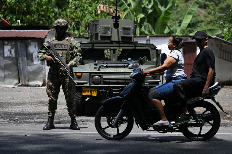 Duas pessoas em uma motocicleta passam por um homem em uniforme militar que carrega uma arma grande.