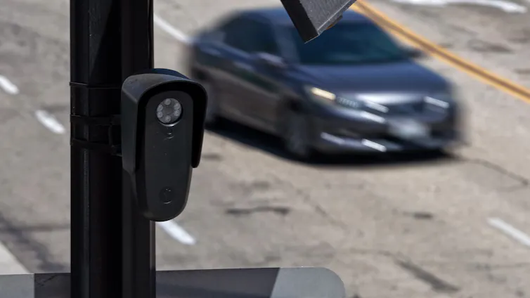 a vertical black box with a translucent circle near the top on a pole beside a road as a car passes by