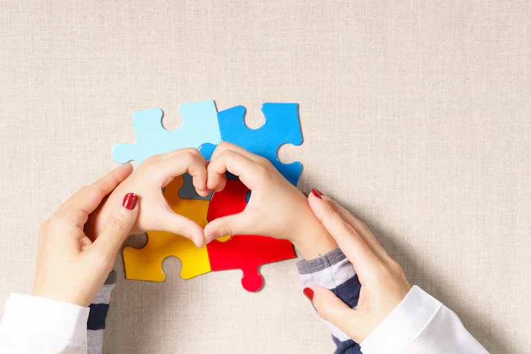 Top view of adult hands around the hands of a small child making heart shape over four brightly coloured jigsaw pieces.