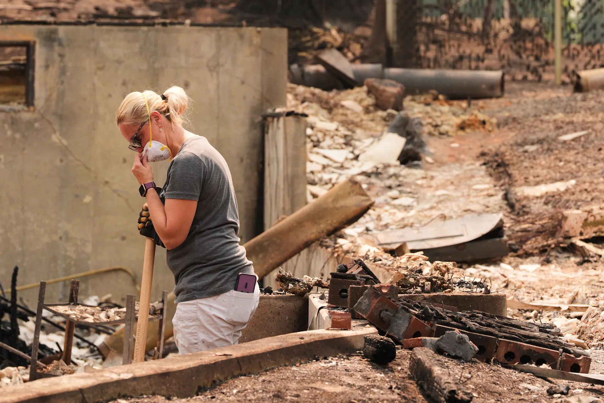 A woman wearing a construction mask stands amid the ash and debris of a home.