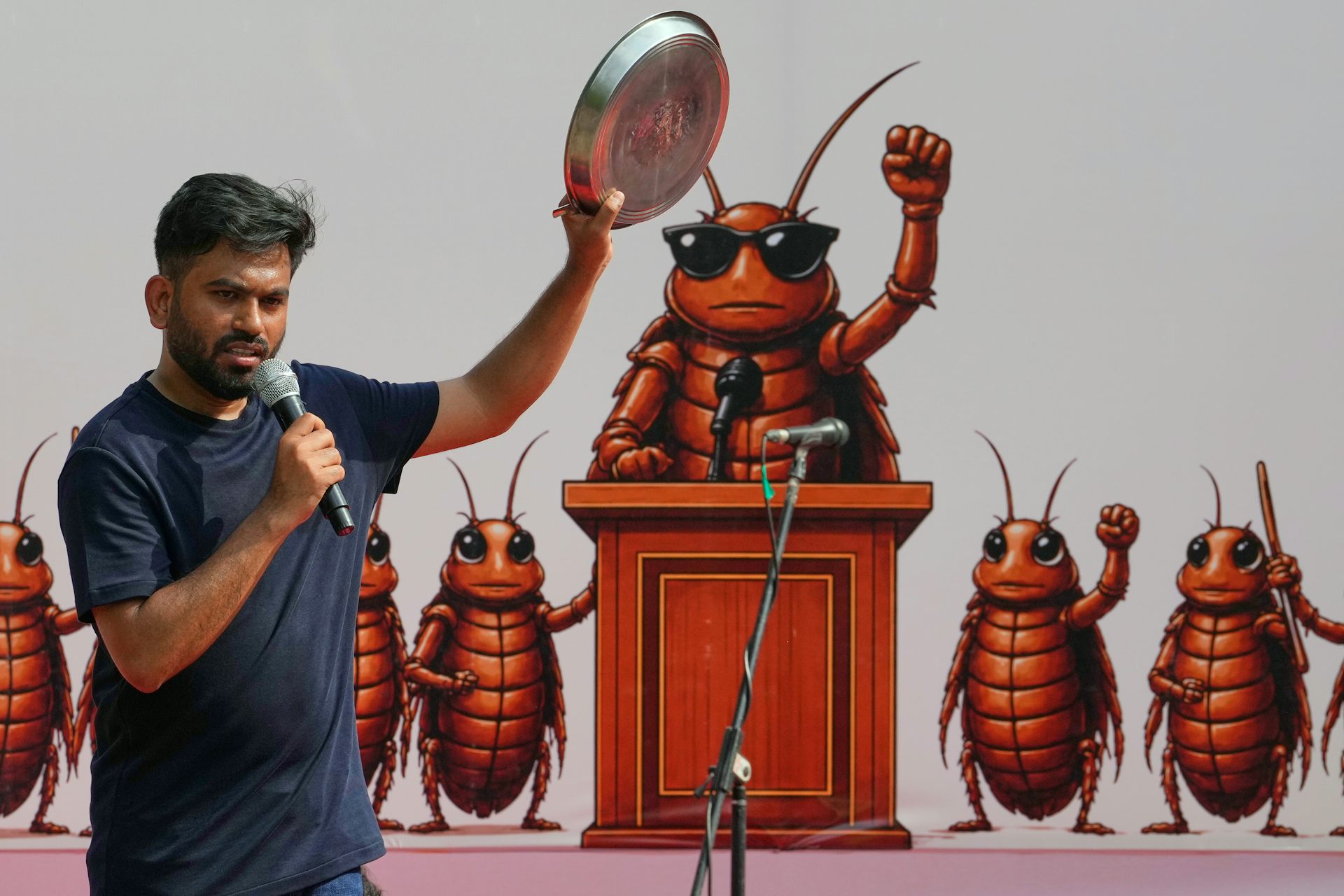 A dark-haired man with a beard speaks into a microphone in front of a mural showing cartoon cockroaches.