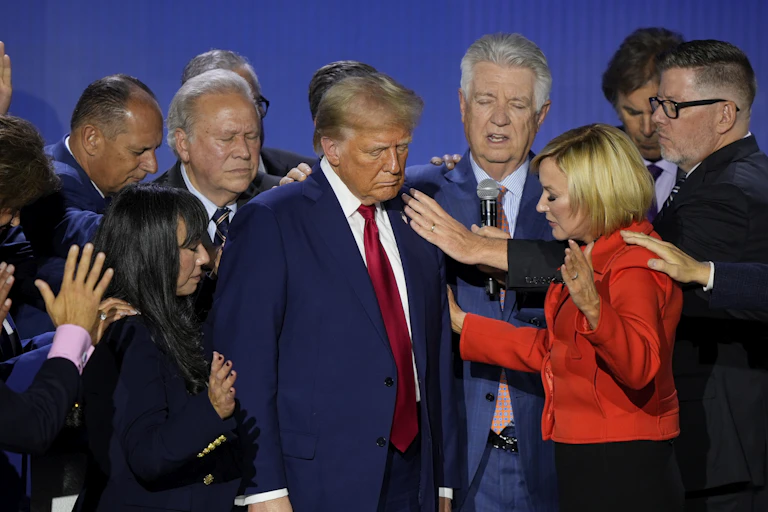 A hulking man with blondish-orange fluffy hair stands with his eyes shut as people pray around him,