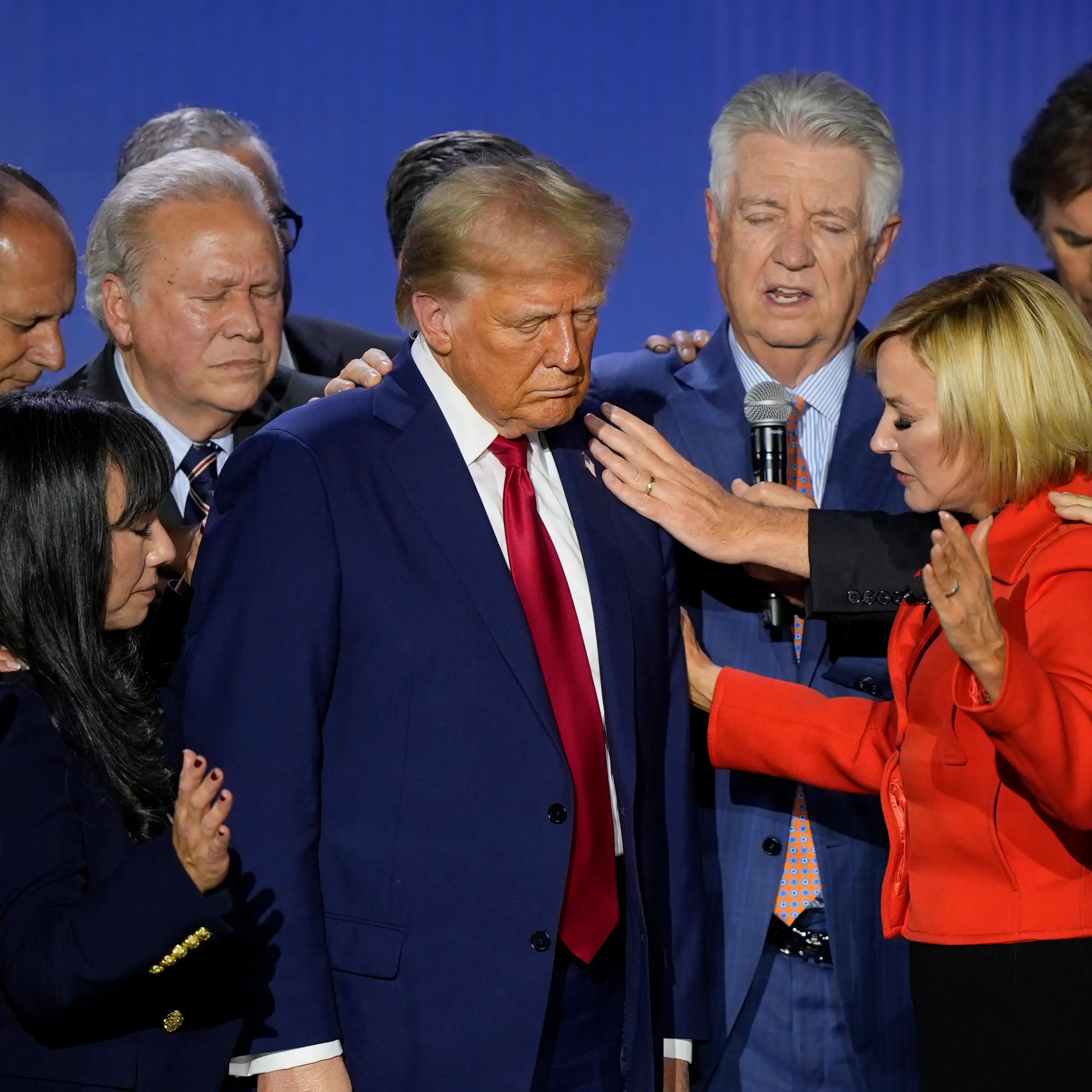 A hulking man with blondish-orange fluffy hair stands with his eyes shut as people pray around him,