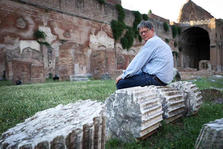 A middle-aged man poses while sitting on a toppled Roman column surrounded by grass and ruins.