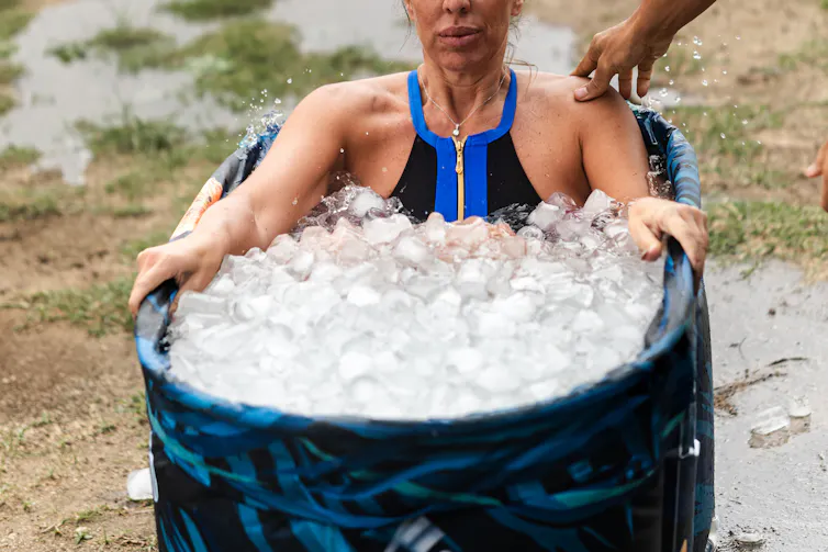 A muscular woman in a workout top sits in an ice bath