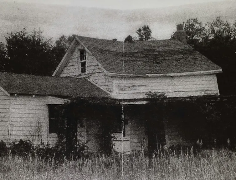 A black-and-white photo of a two-story white house with a one-story section on one end.