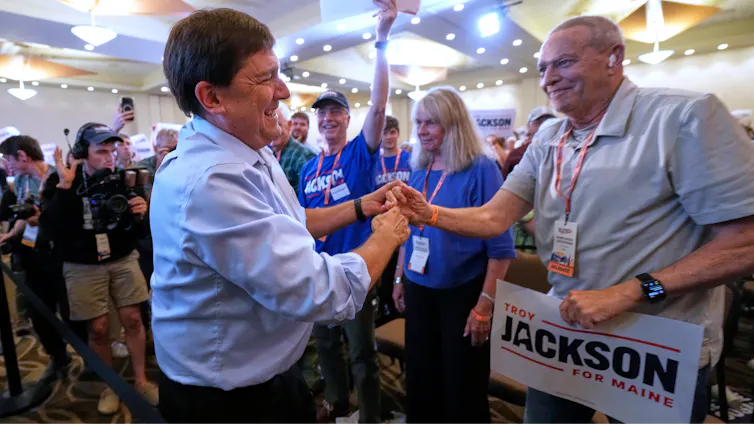 A smiling man clasps hands with another man holding a 'Troy Jackson' campaign sign at a political rally.