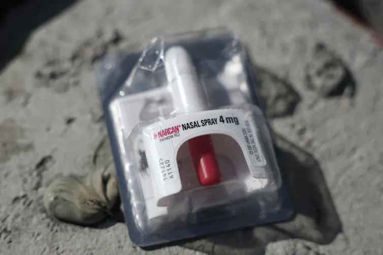 A white and red nasal spray container is wrapped in plastic and rests on a grey pillar.