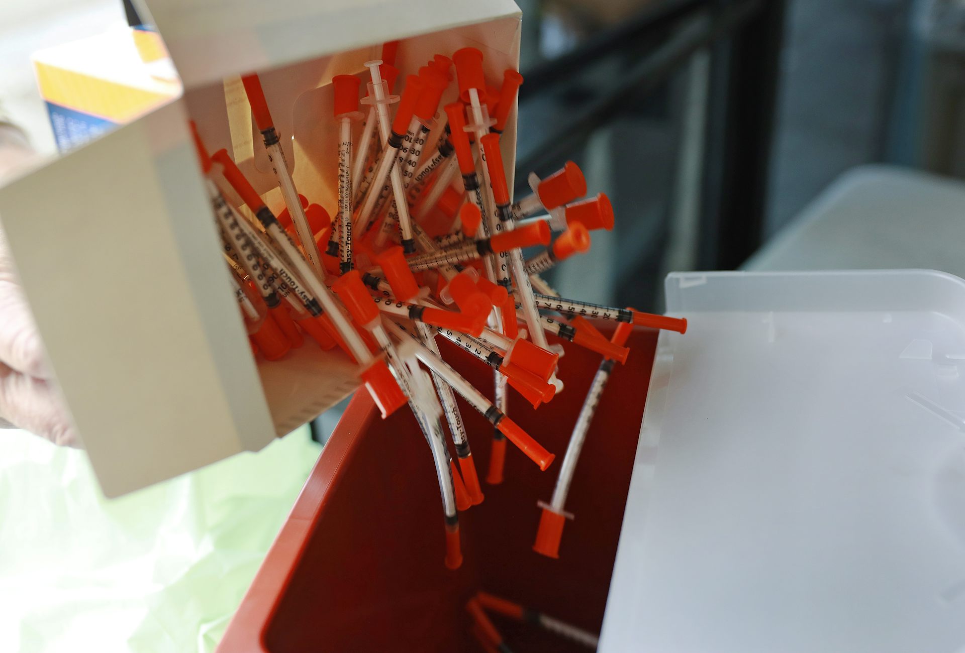A white box releases many needles with an orange backing into a red square container.