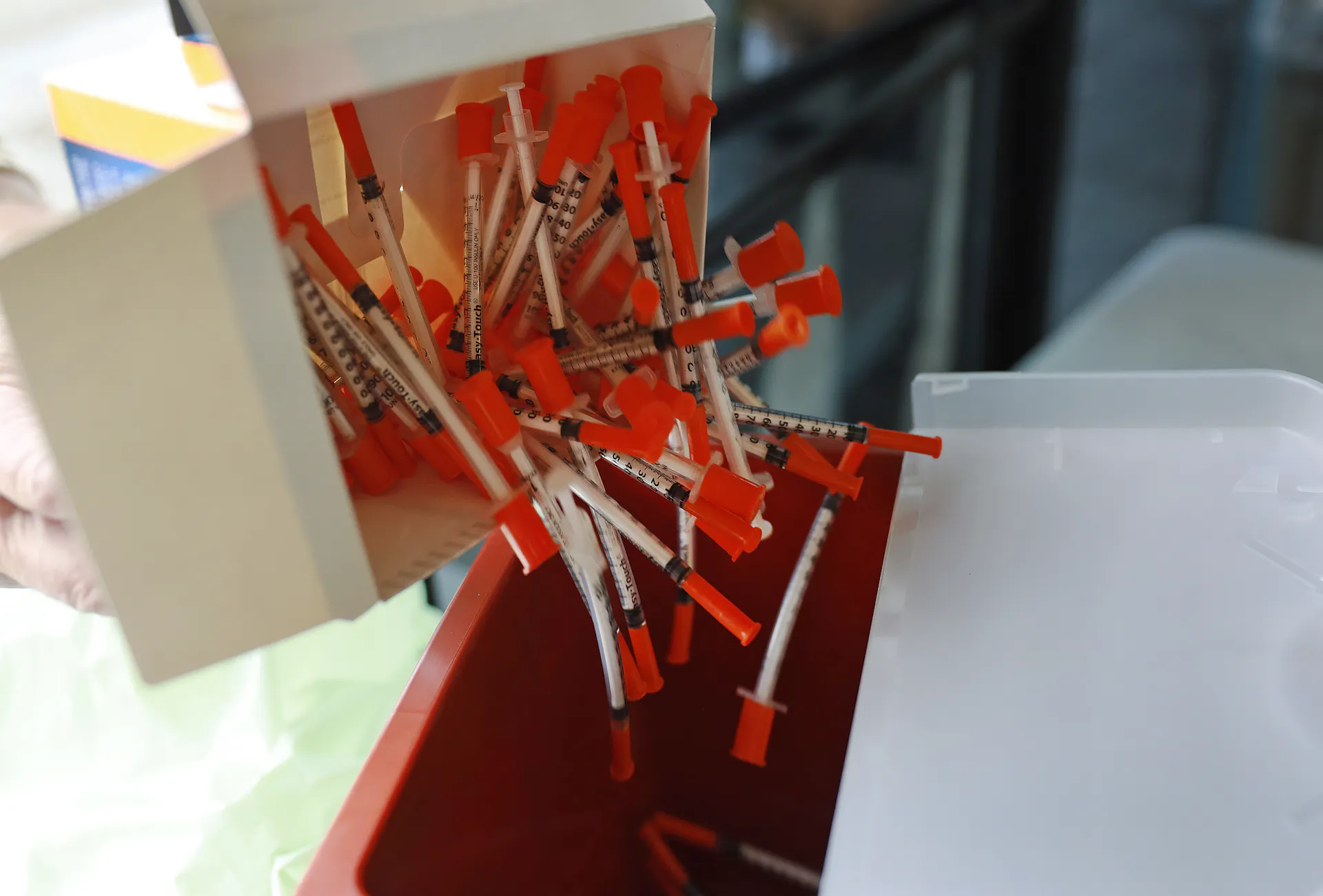 A white box releases many needles with an orange backing into a red square container.