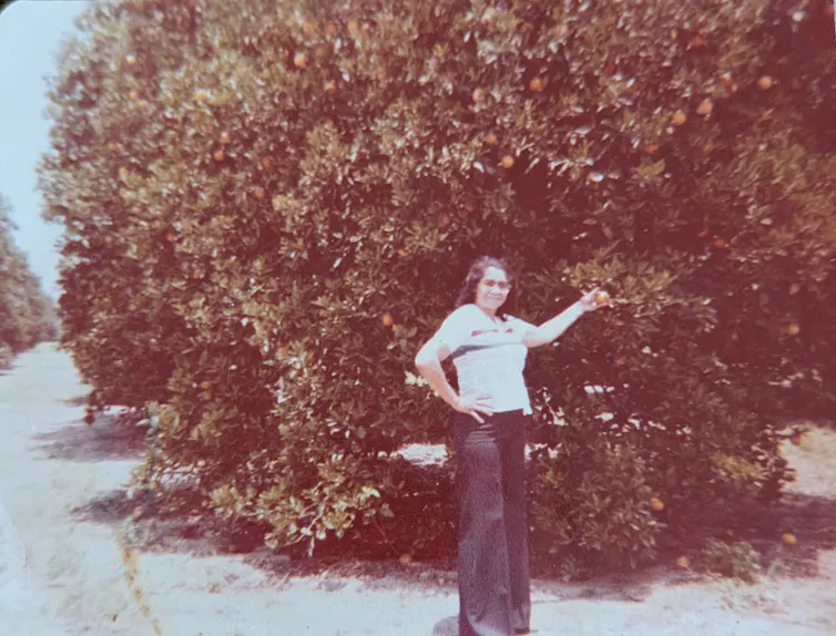 A person in a white shirt and dark pants stands next to a tree.