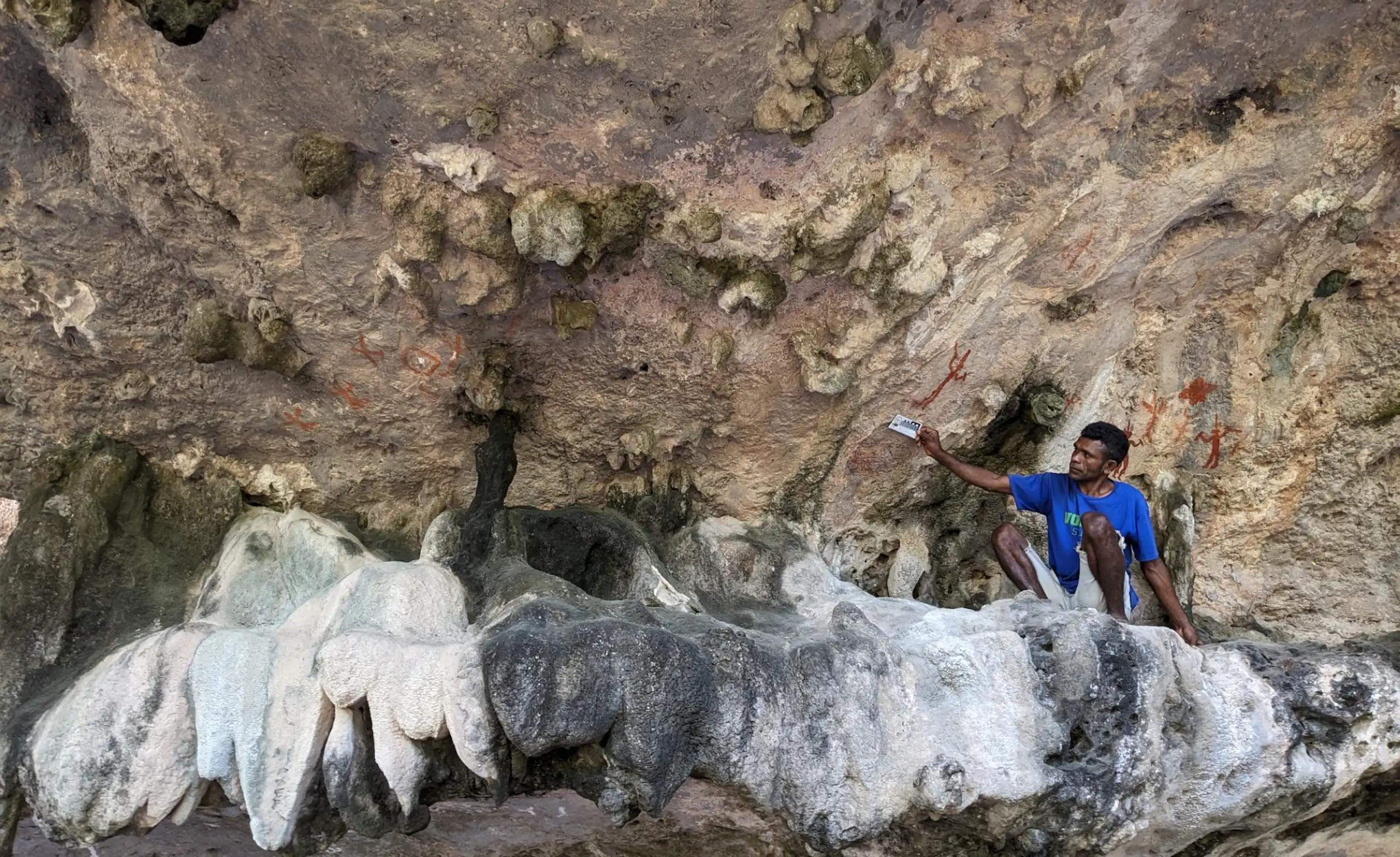 A man sits underneath what looks like a rocky outcrop, with red pigment visible on the walls behind.