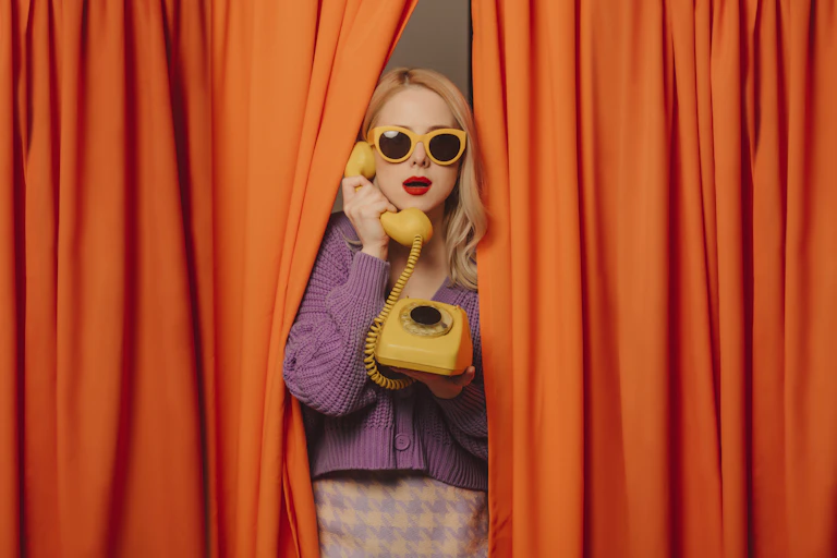 A woman in a retro outfit stands among orange curtains with a yellow rotary phone.