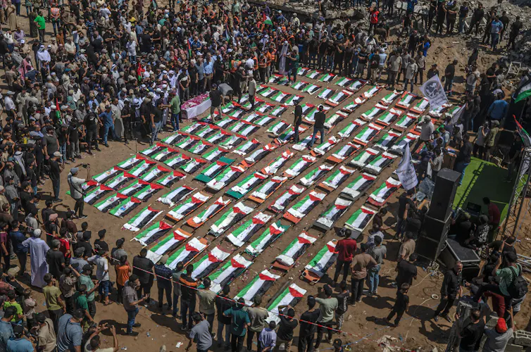 A large crowd of people around the coffins of 112 people which are wrapped in Palestinian flags.