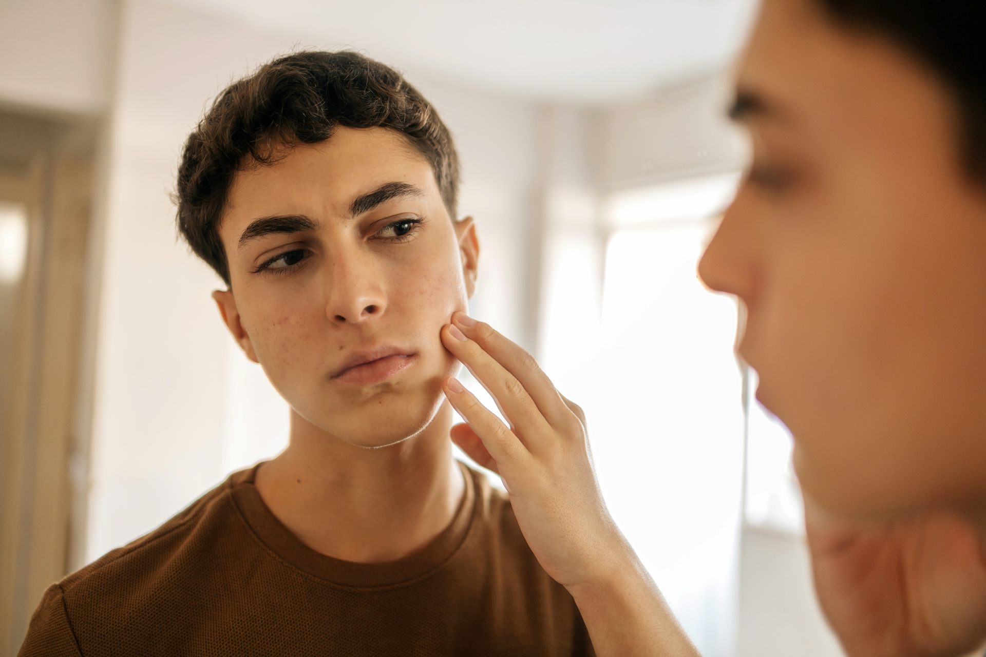 A teen boy looks at himself in the mirror while touching his face. He looks concerned or self conscious.