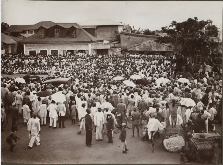 A vintage black and white photo of a large crowd of smartly dressed Nigerians in a town square.