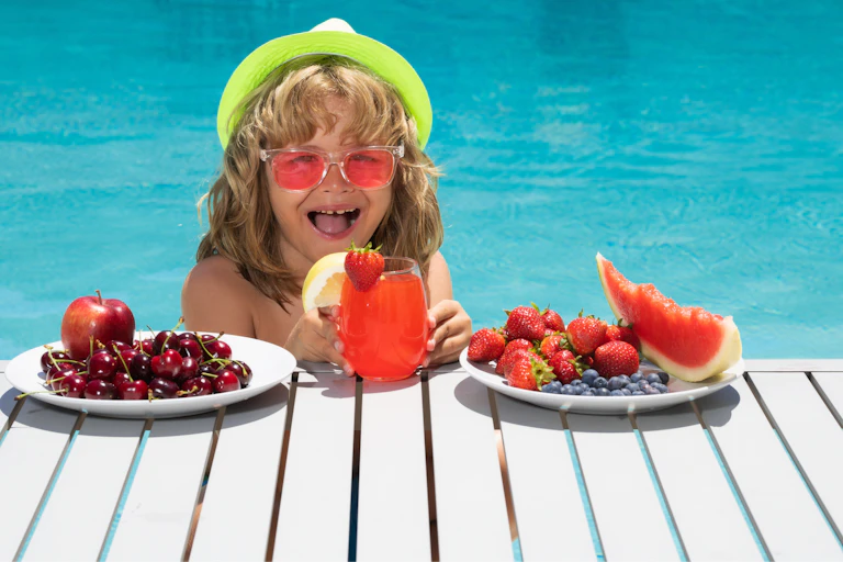 Happy looking child wearing brightly coloured sunglasses and hat is at edge of outdoor swimming pool, where there are two plates of red coloured berries and fruits and a glass of red fluid with fruit embellishments