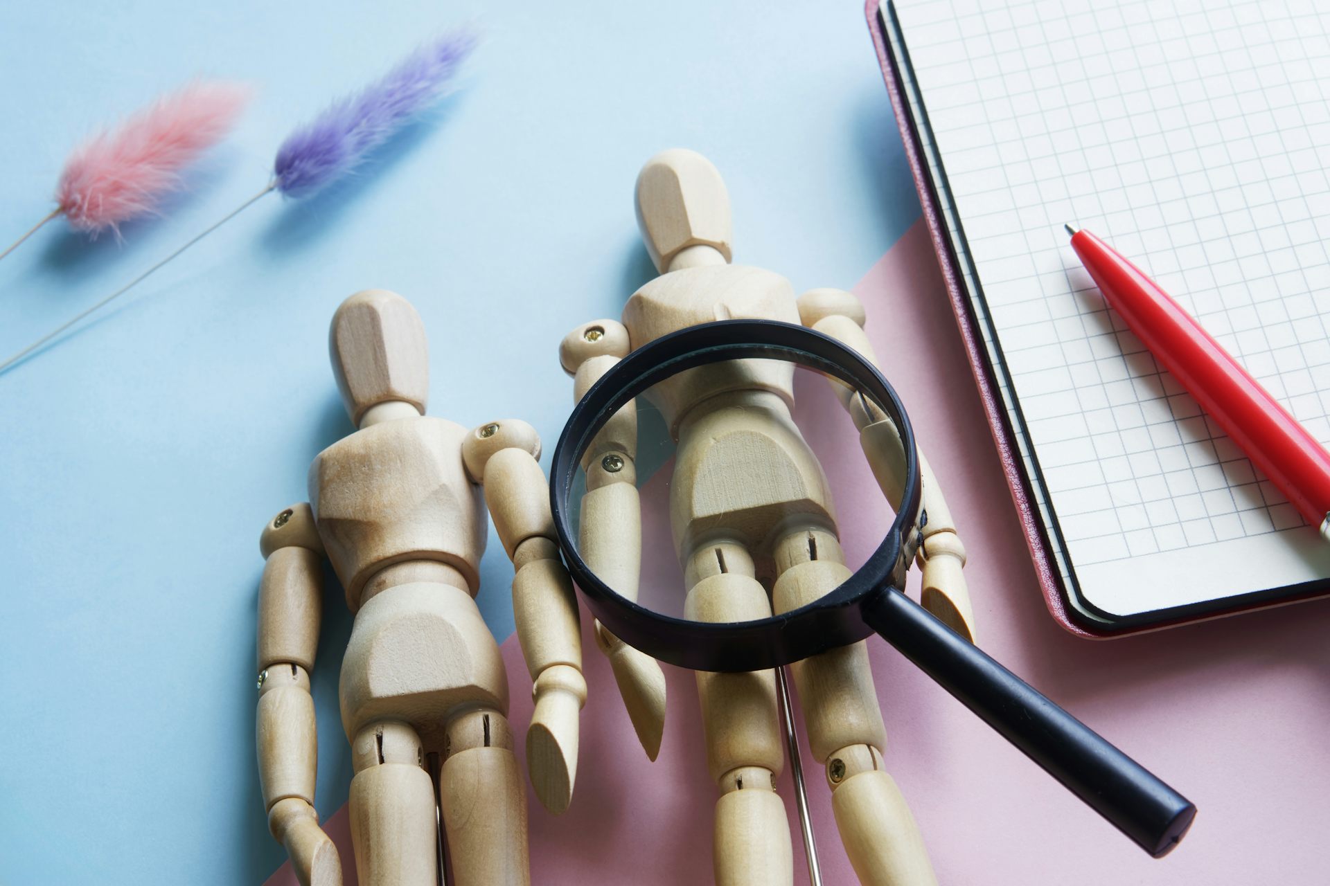Small wooden mannequins with magnifying glass against pink and blue background.
