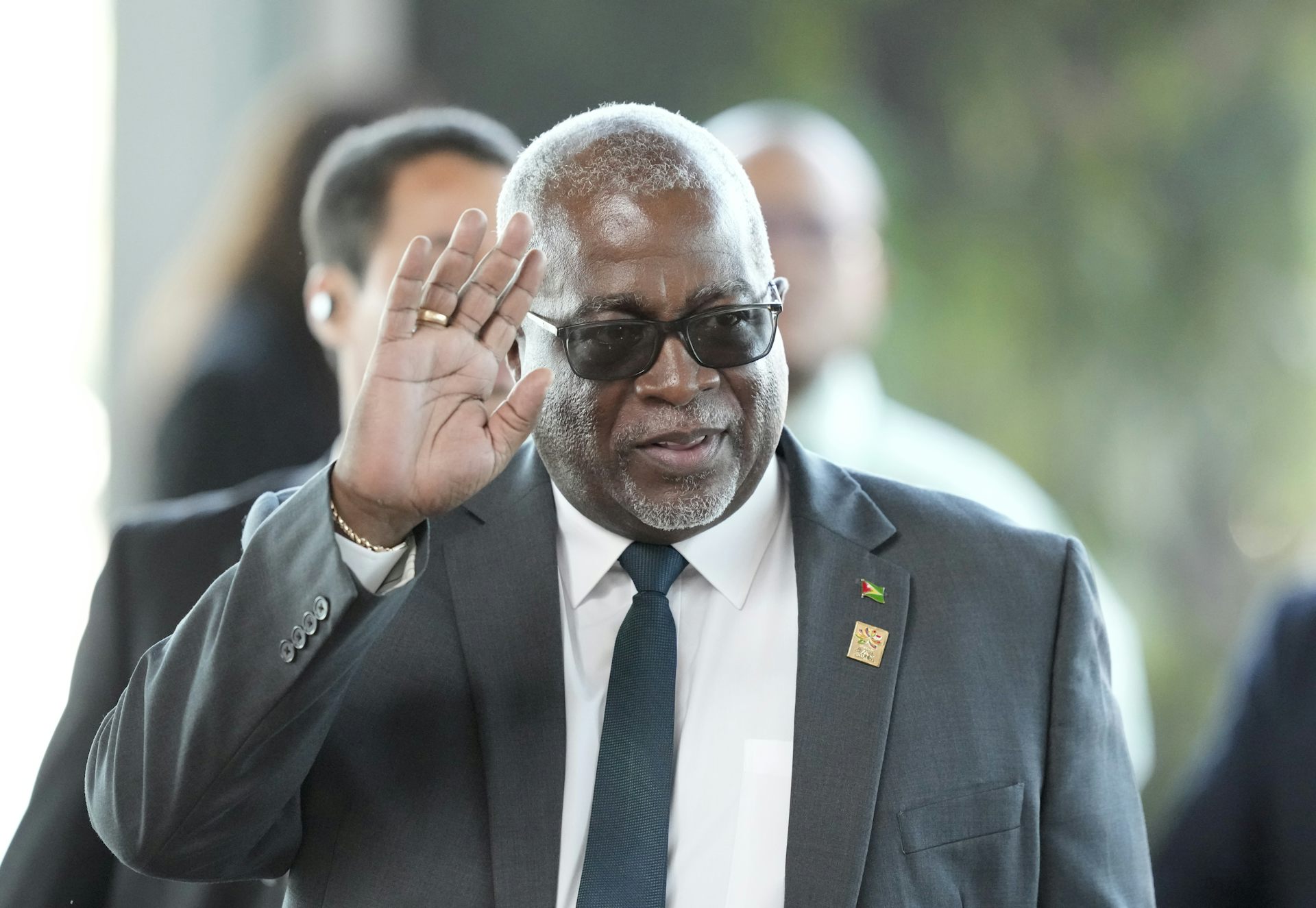 A Black man in a suit and sunglasses waves.