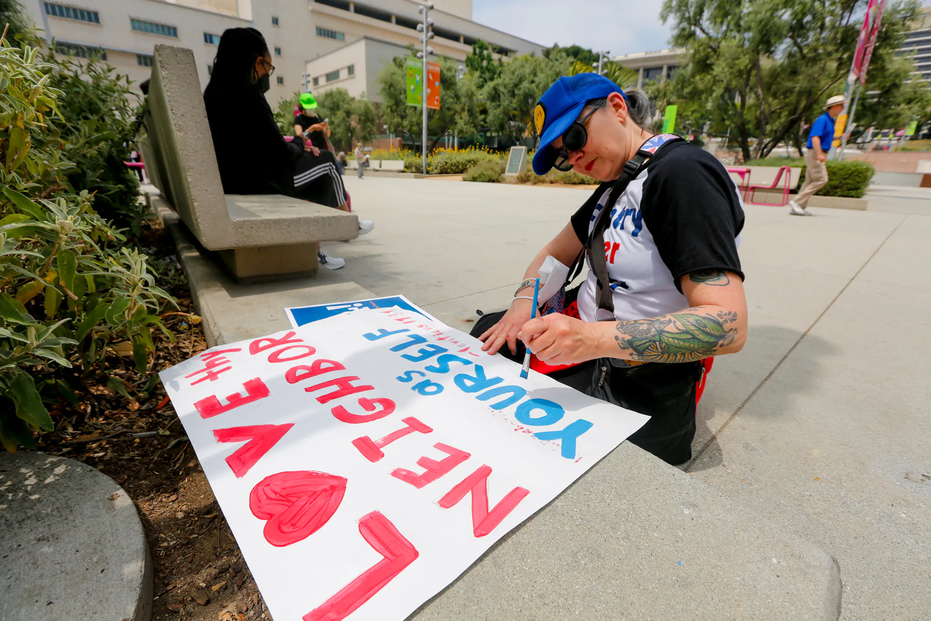 A woman seated on the ground writes, 'Love thy neighbor as yourself' on a poster.