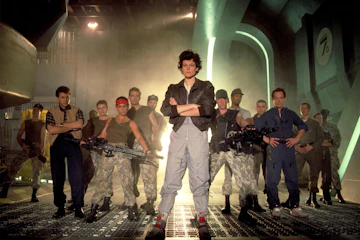 The cast of Aliens stand together with Sigourney Weaver at the front.