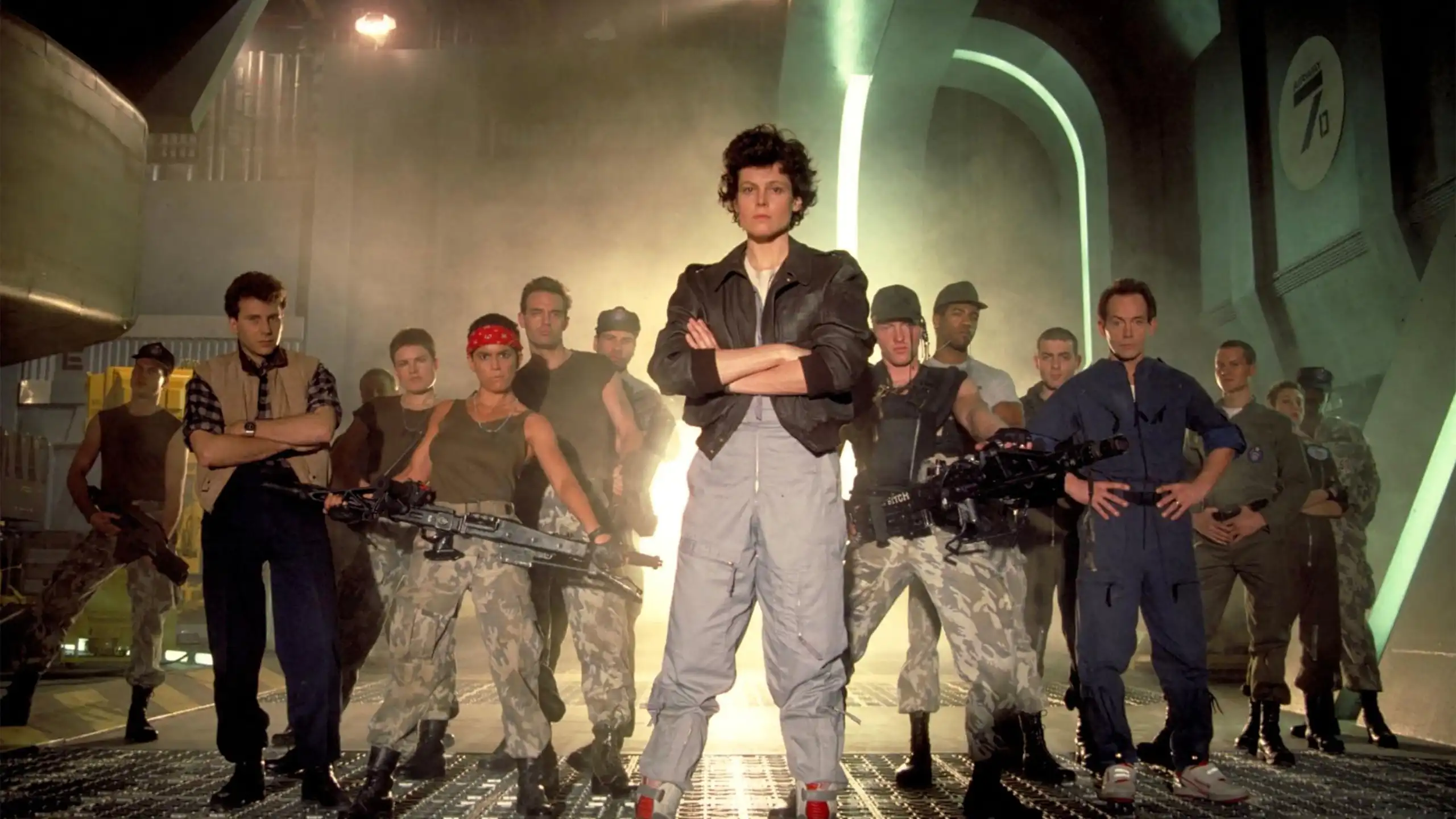 The cast of Aliens stand together with Sigourney Weaver at the front.