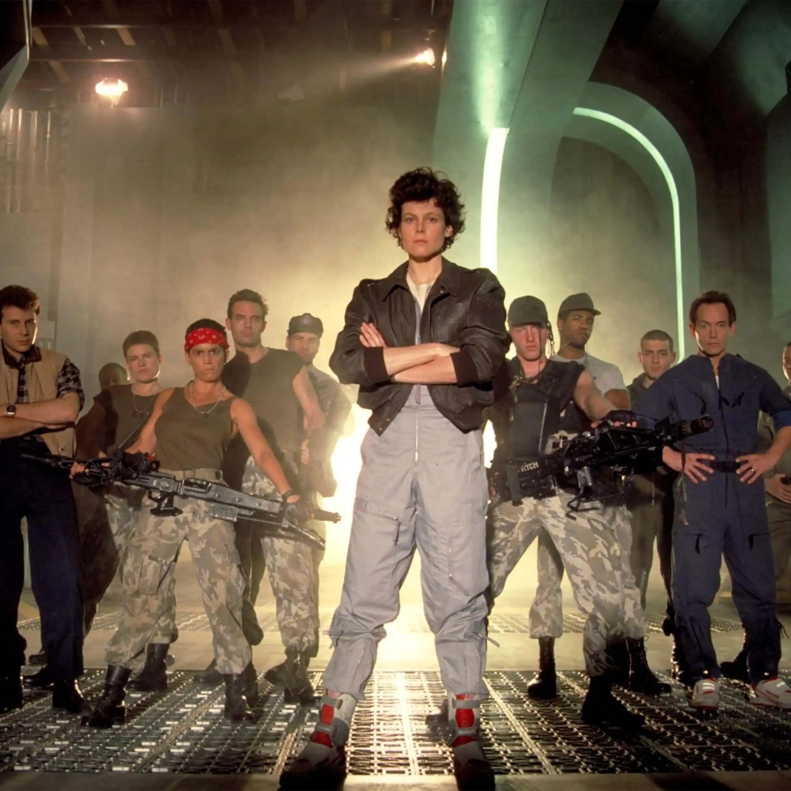 The cast of Aliens stand together with Sigourney Weaver at the front.