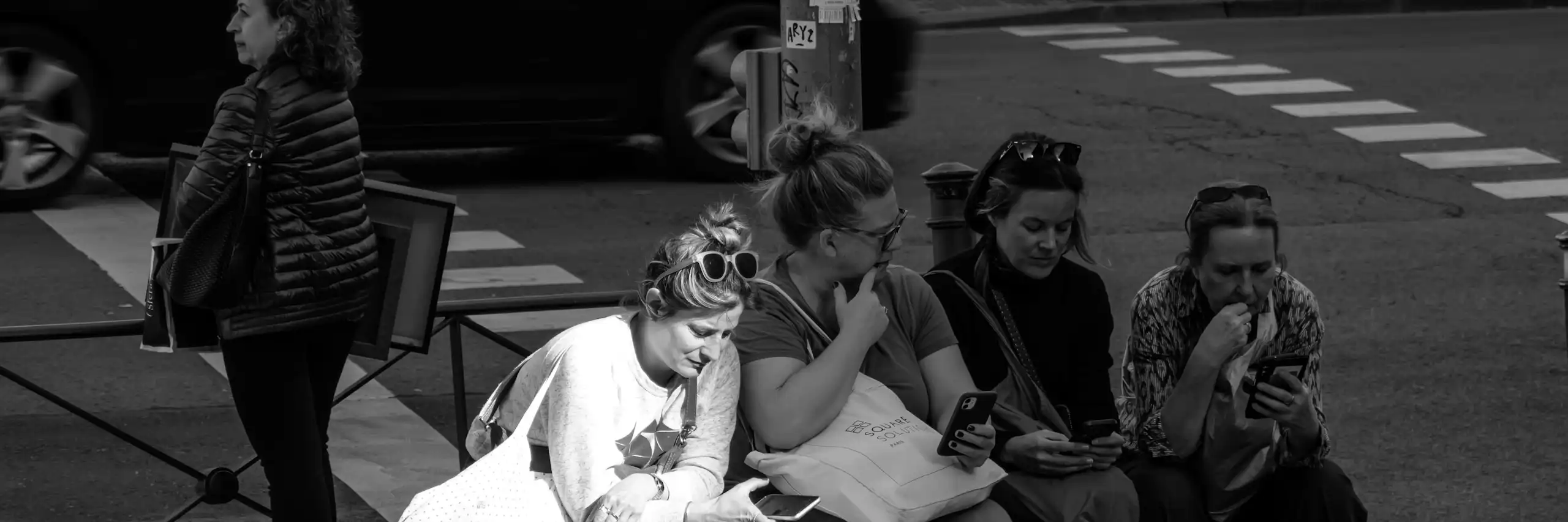Cliché noir et blanc, des jeunes femmes assises sur un banc, chacune un smartphone à la main, discutant tout en cherchant sur leur mobile.