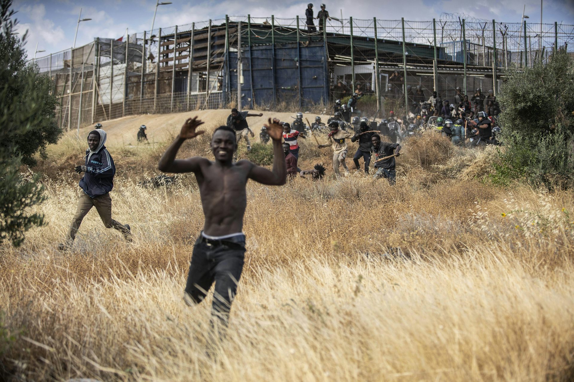 A smiling Black man runs through a grassy field with a fortified fence behind him.