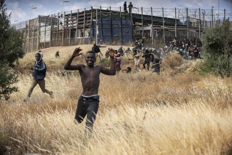 A smiling Black man runs through a grassy field with a fortified fence behind him.