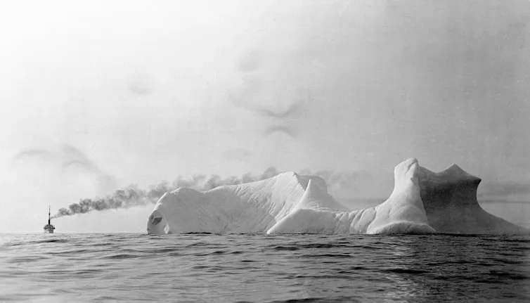 A large iceberg sits in the middle of a large body of water. A ship emitting a plume of smoke can be seen in the distance.