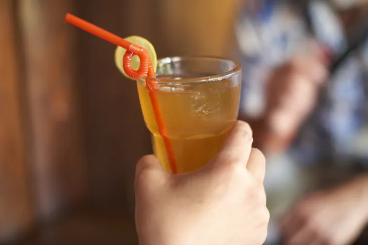 A hand grips a clear glass of orange liquid with ice, featuring a slice of lemon on the rim and a red curly straw.