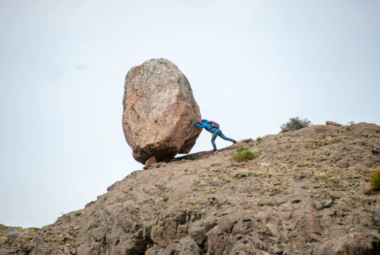 A hiker pushes a large boulder balancing on top of a rocky hill.