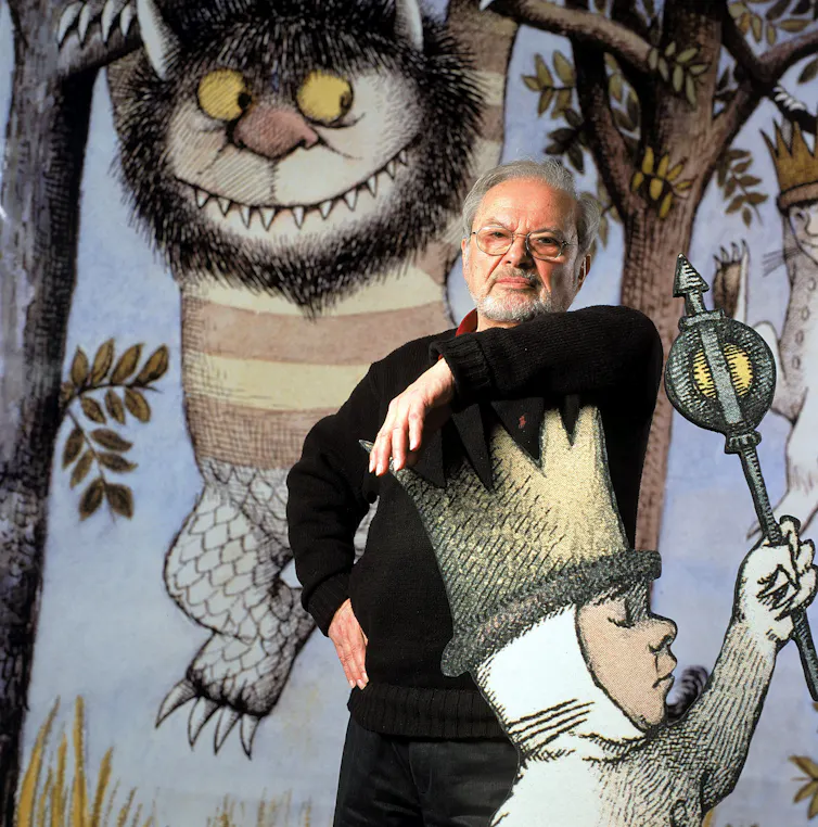 Un homme se lève et pose son bras contre la couronne d'un grand enfant de dessin animé découpé. Des images de monstres de dessins animés suspendus aux arbres sont visibles derrière lui.