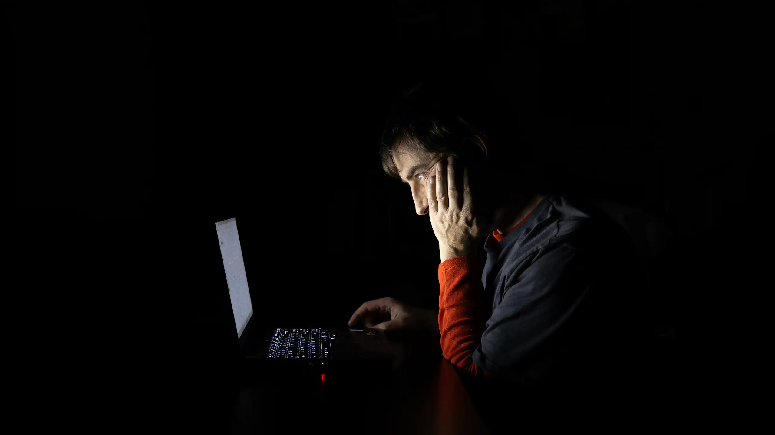Um jovem, na penumbra, olha para a tela de um laptop