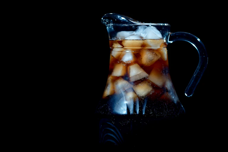 A glass pitcher with dark fluid and lots of ice cubes against a black background