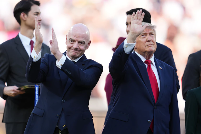 two old men in dark suits, one bald, one orange-skinned, wave to a crowd