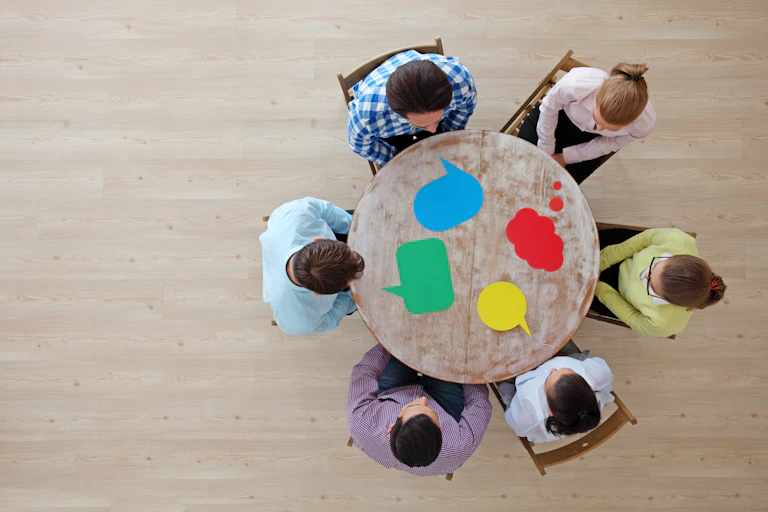 Seen from above, a group of six people sit around a table with different color speech bubbles in the middle.