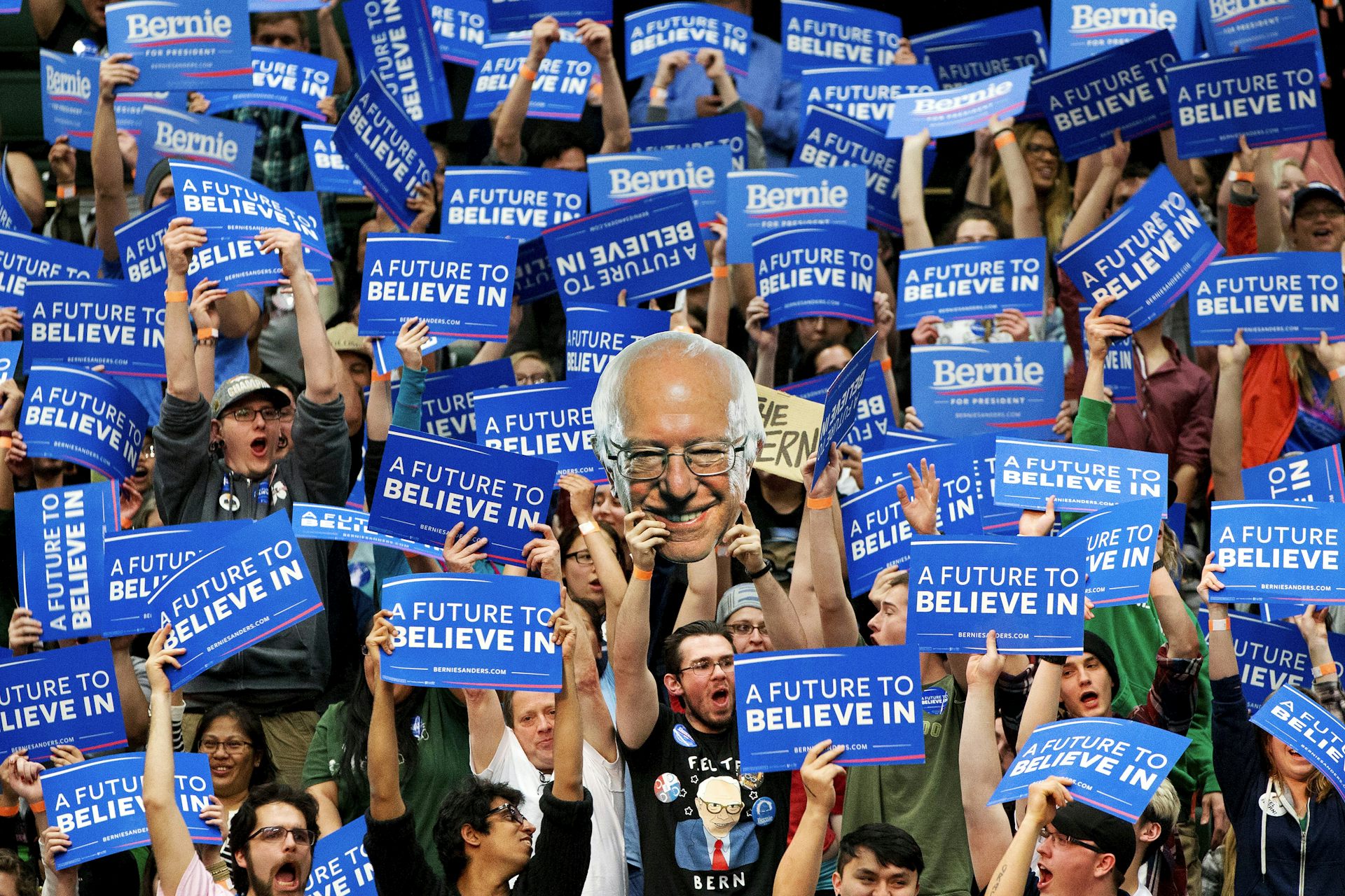 Young people holding signs supporting Bernie Sanders, including a cutout of Sanders' head.