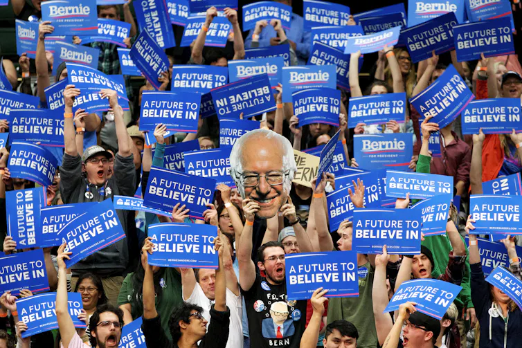 Young people holding signs supporting Bernie Sanders, including a cutout of Sanders' head.
