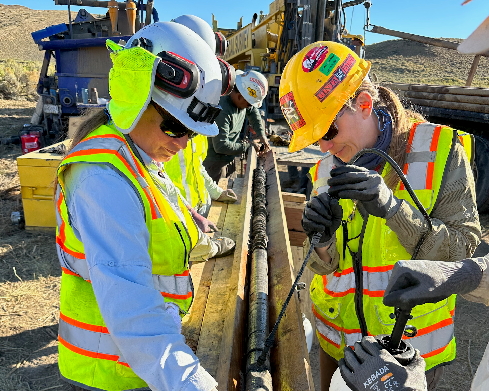 Two scientists in hard hats, with heavy machinery in the background. One scientists is separating part of a long cylinder of mud and sediment.