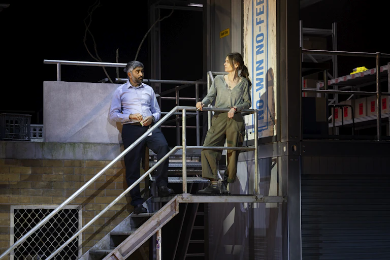 Production image: a man and a woman stand on a metal staircase.