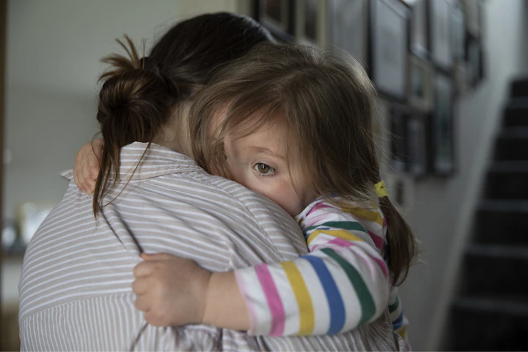 Young girl gripping tightly to her mother in a reassuring embrace, with her arms wrapped around her mother's shoulders, in a domestic setting.
