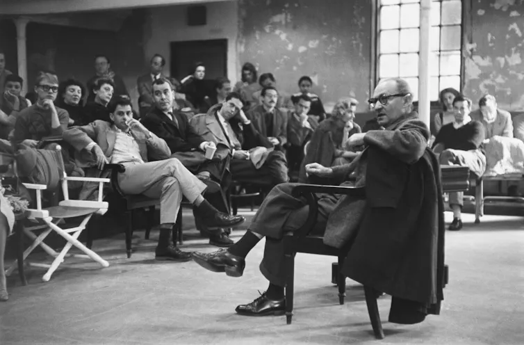 Black and white photo: Stasberg in a chair, students in the background.