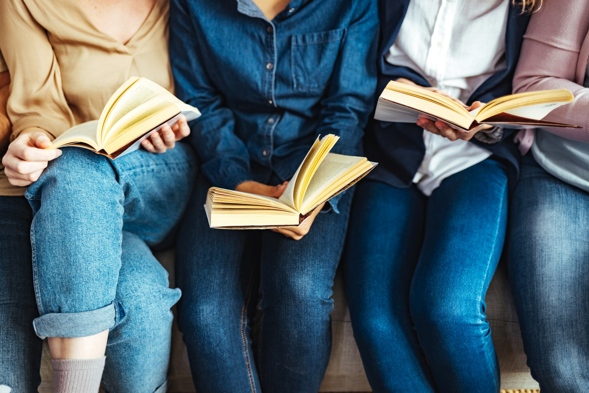 Diverse group of friends discussing a book in a library. No faces visible, all wear jeans.