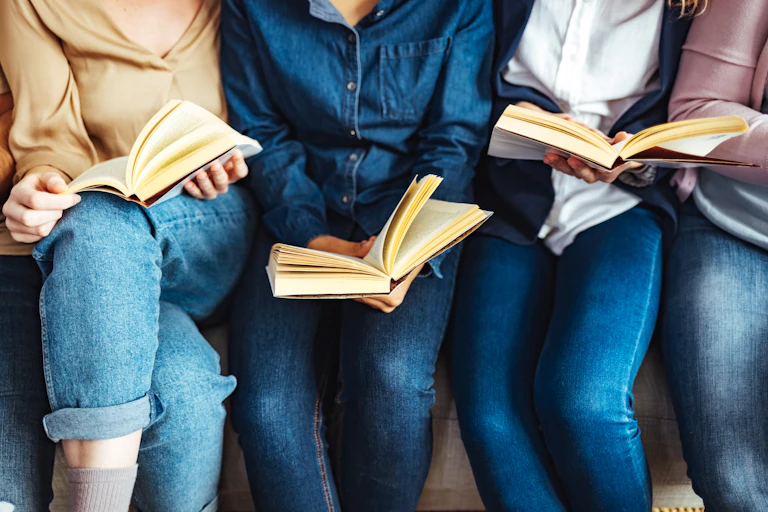 Diverse group of friends discussing a book in a library. No faces visible, all wear jeans.
