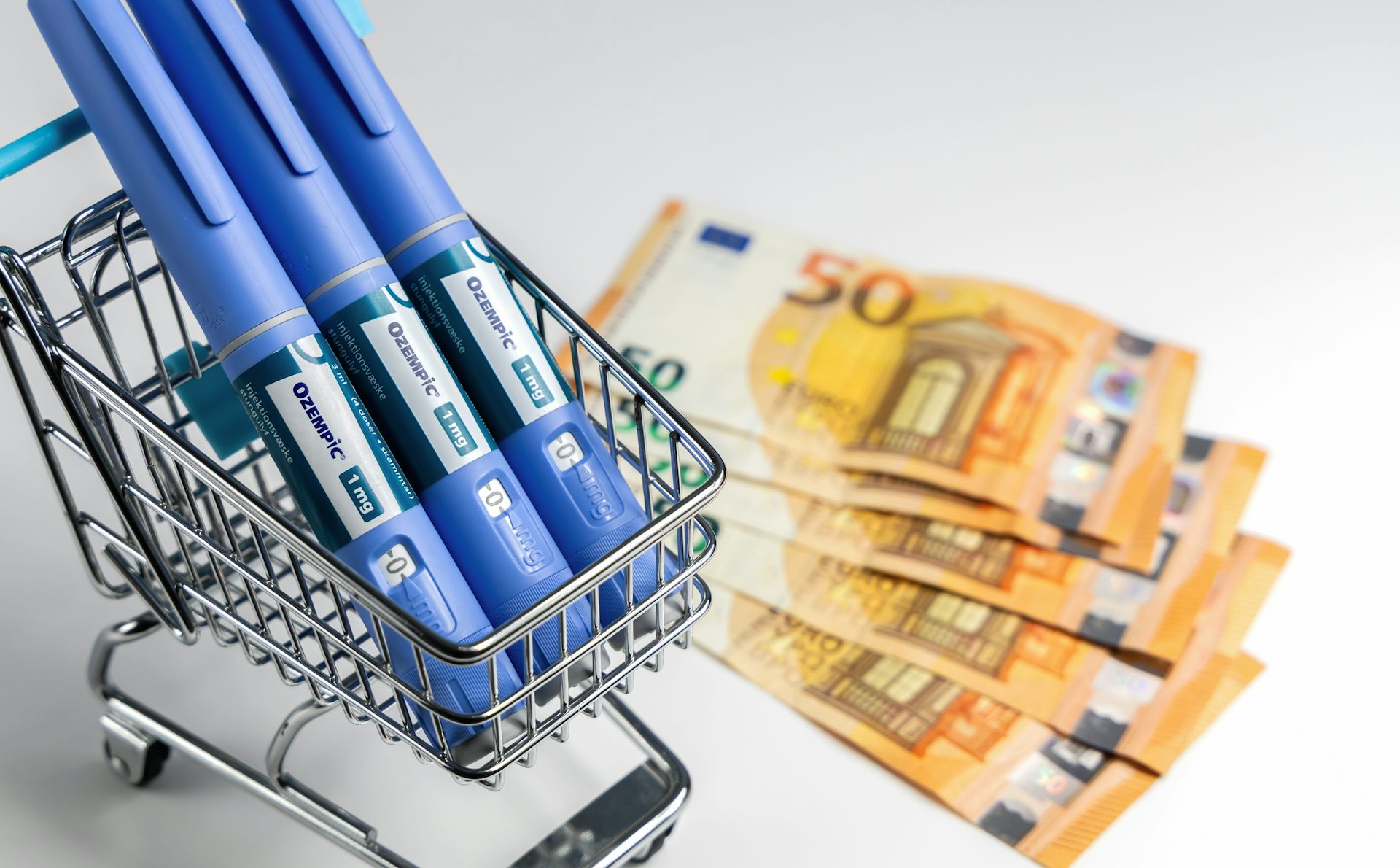 Three Ozempic pens in a miniature supermarket trolley with cash notes next to them. 